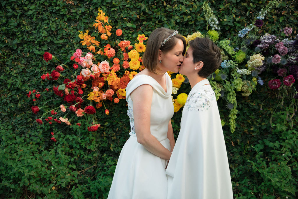 brides in coordinating bridal dress and bridal jumpsuit kiss in front of hanging rainbow floral installation after wedding ceremony at The Fig House