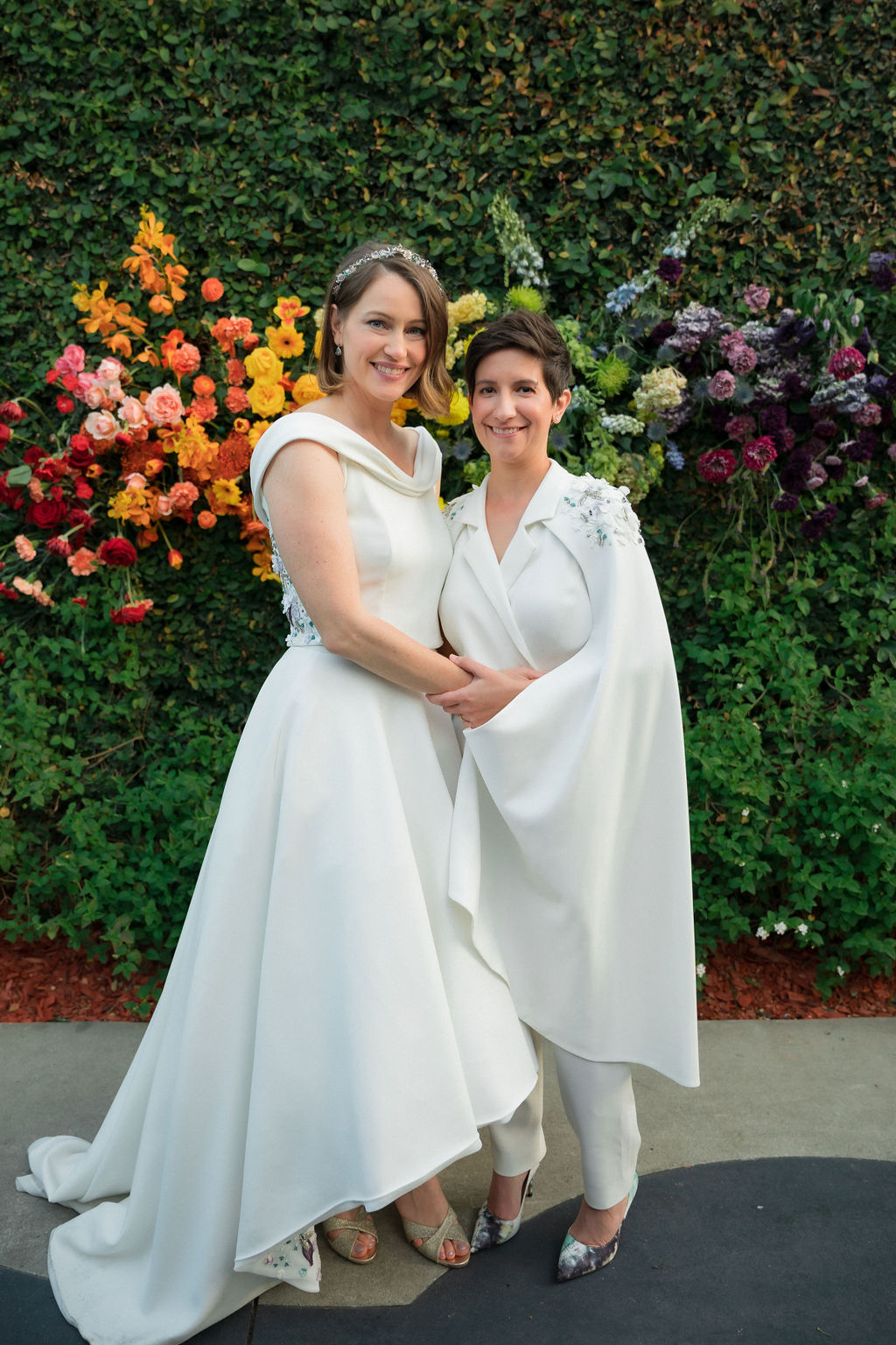 brides in coordinating bridal dress and bridal jumpsuit embrace in front of hanging rainbow floral installation after wedding ceremony at The Fig House