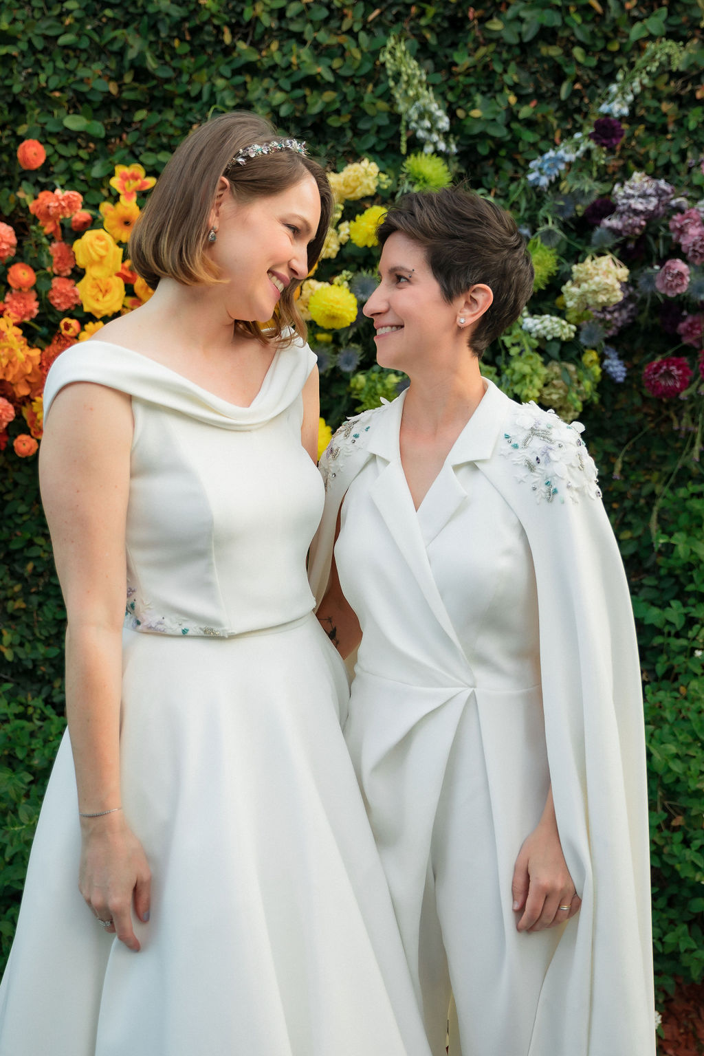 brides in coordinating bridal dress and bridal jumpsuit look at one another in front of hanging rainbow floral installation after wedding ceremony at The Fig House