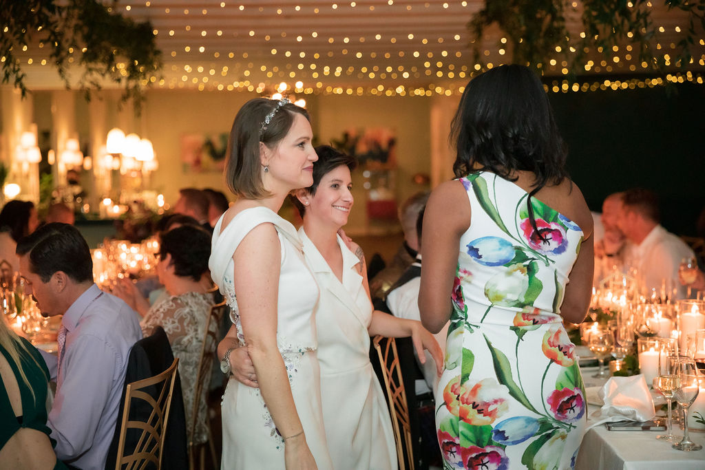 two brides holding each other talk to guest during wedding reception 