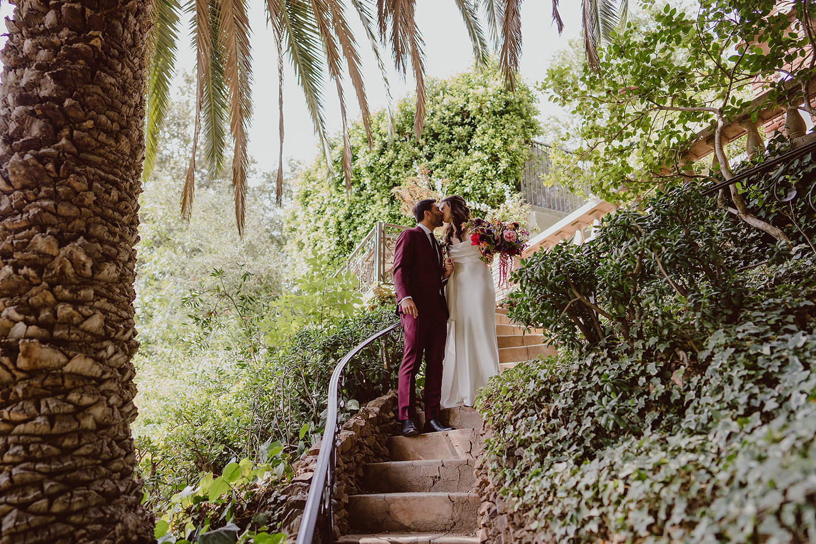 bride in minimalist satin wedding dress stands on staircase with groom in burgundy suit and black tie before their magical wedding at The Houdini Estate in Hollywood