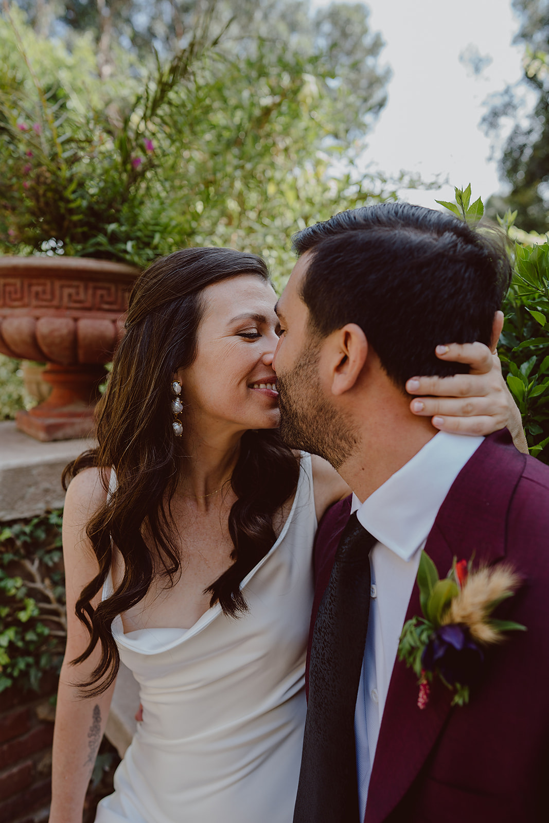minimalist bride in silk dress kisses groom in burgundy suit and black tie before their magical wedding at The Houdini Estate in Hollywood