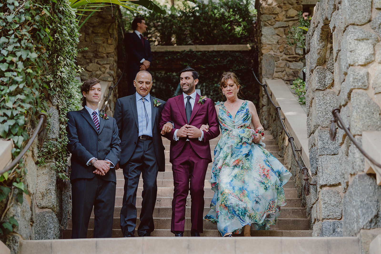 groom in burgundy suit and black tie walks down stair case towards ceremony