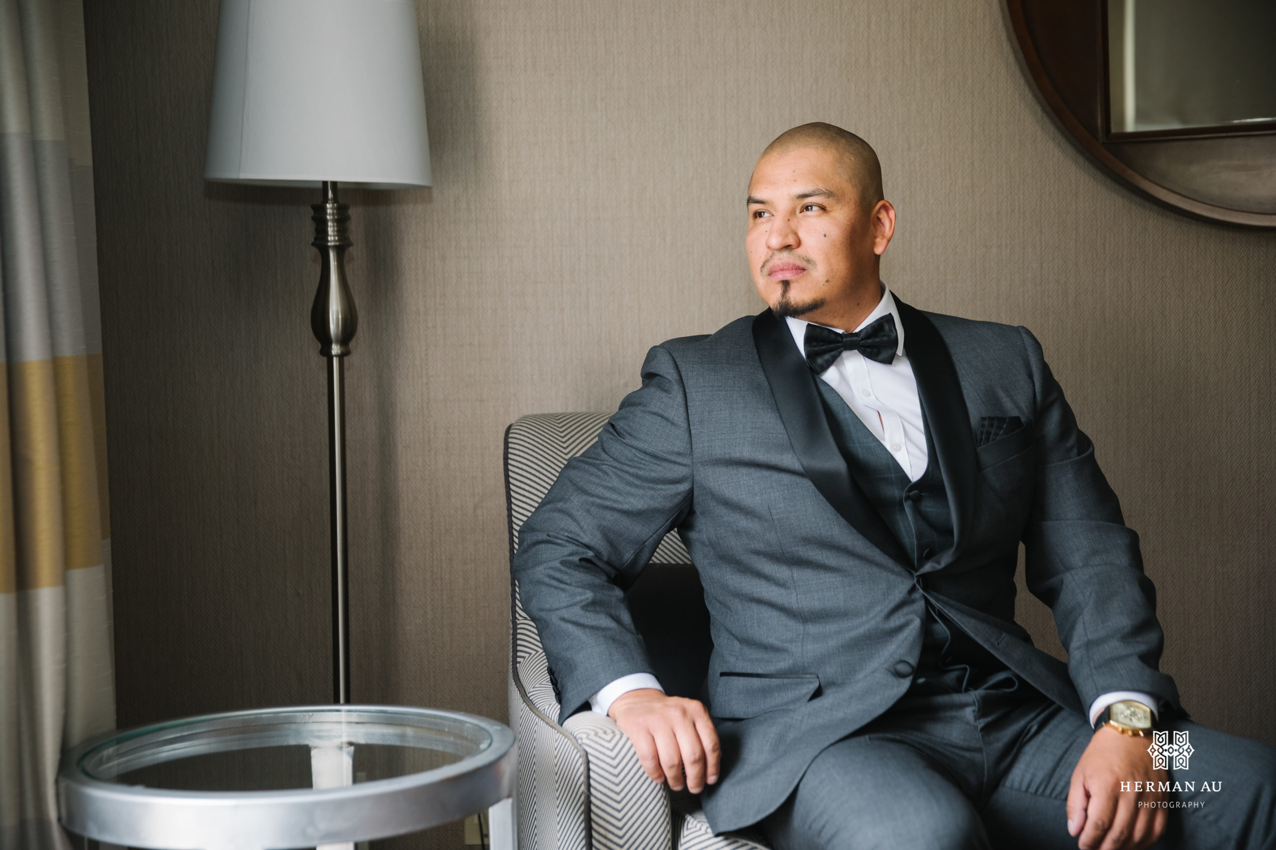 groom in dark grey suit with black bowtie sits in hotel room 