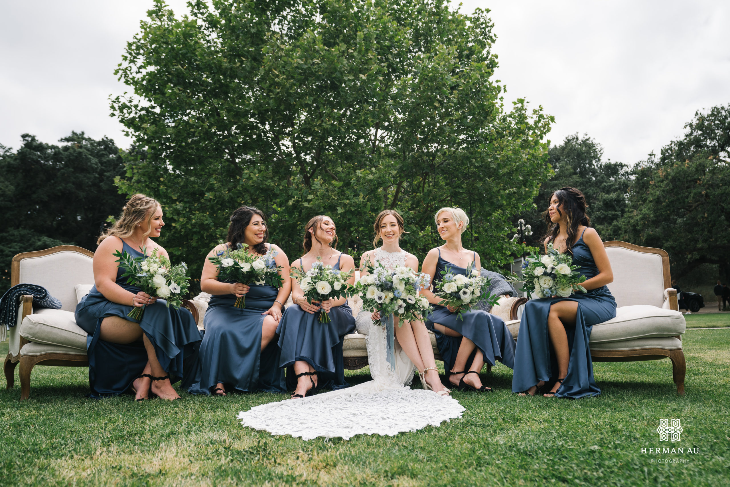  bride in high neckline lace wedding dress and white and blue bridal bouquet sits with bridesmaids in dark blue satin dresses on outdoor couch