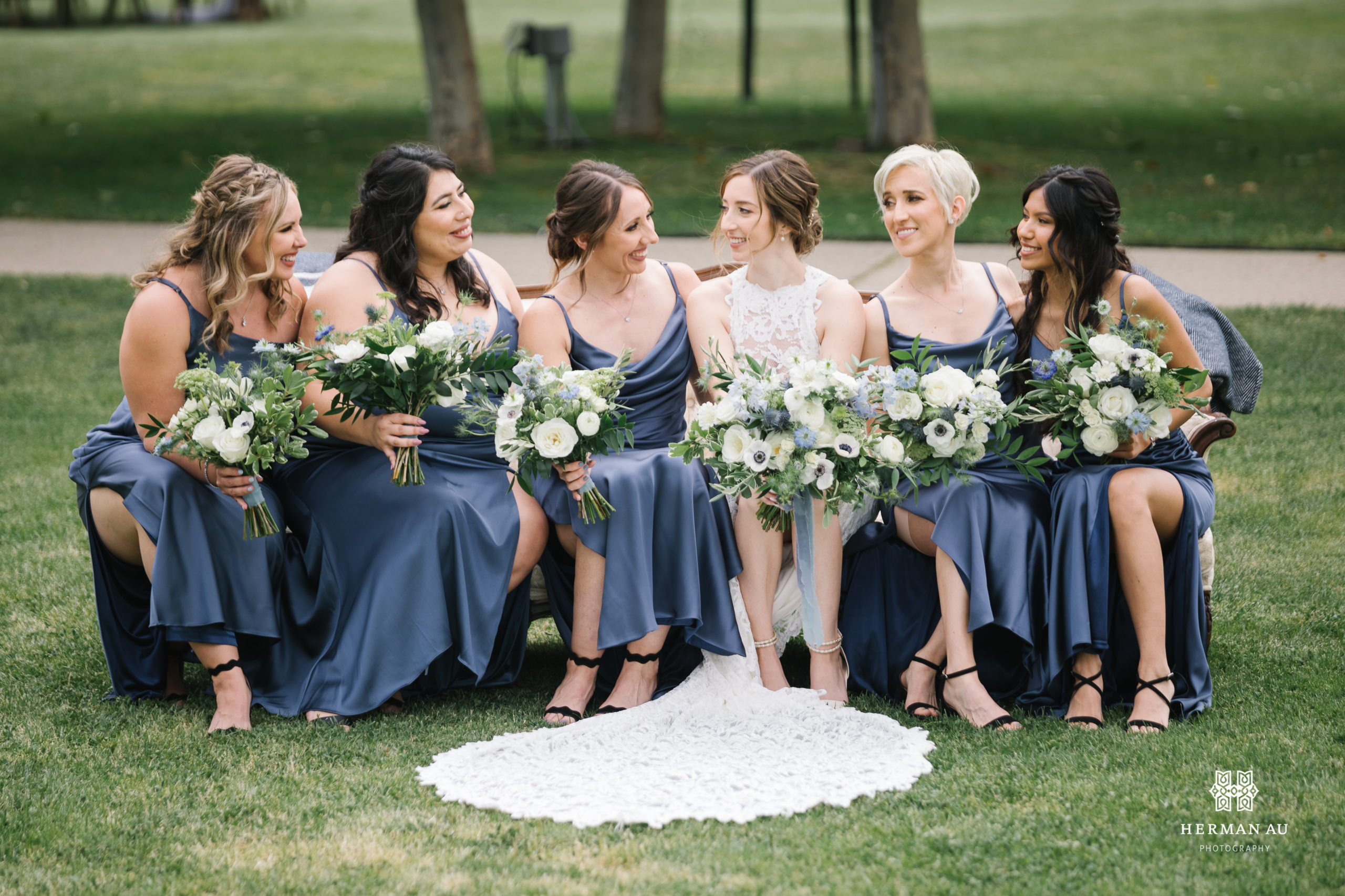 bride in high neckline lace wedding dress and white and blue bridal bouquet sits on couch with bridesmaids in dark blue satin dresses