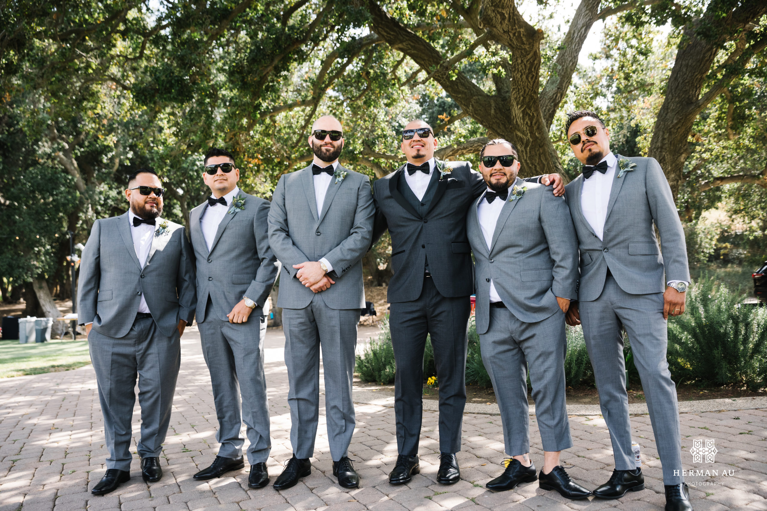groom in dark grey suit and black bowtie with sunglasses stands with groomsmen in light grey suits and black bowties 