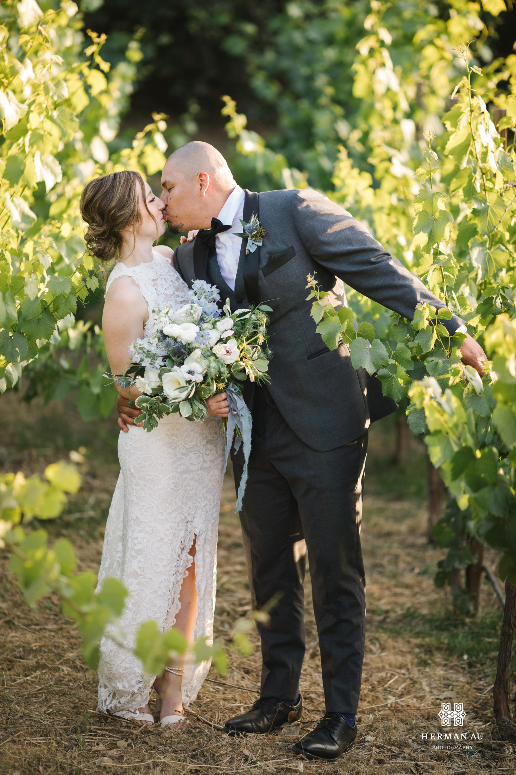 bride in high neckline open back lace wedding dress and groom in dark grey suit and black bowtie take photos in vineyard at Saddlerock Ranch