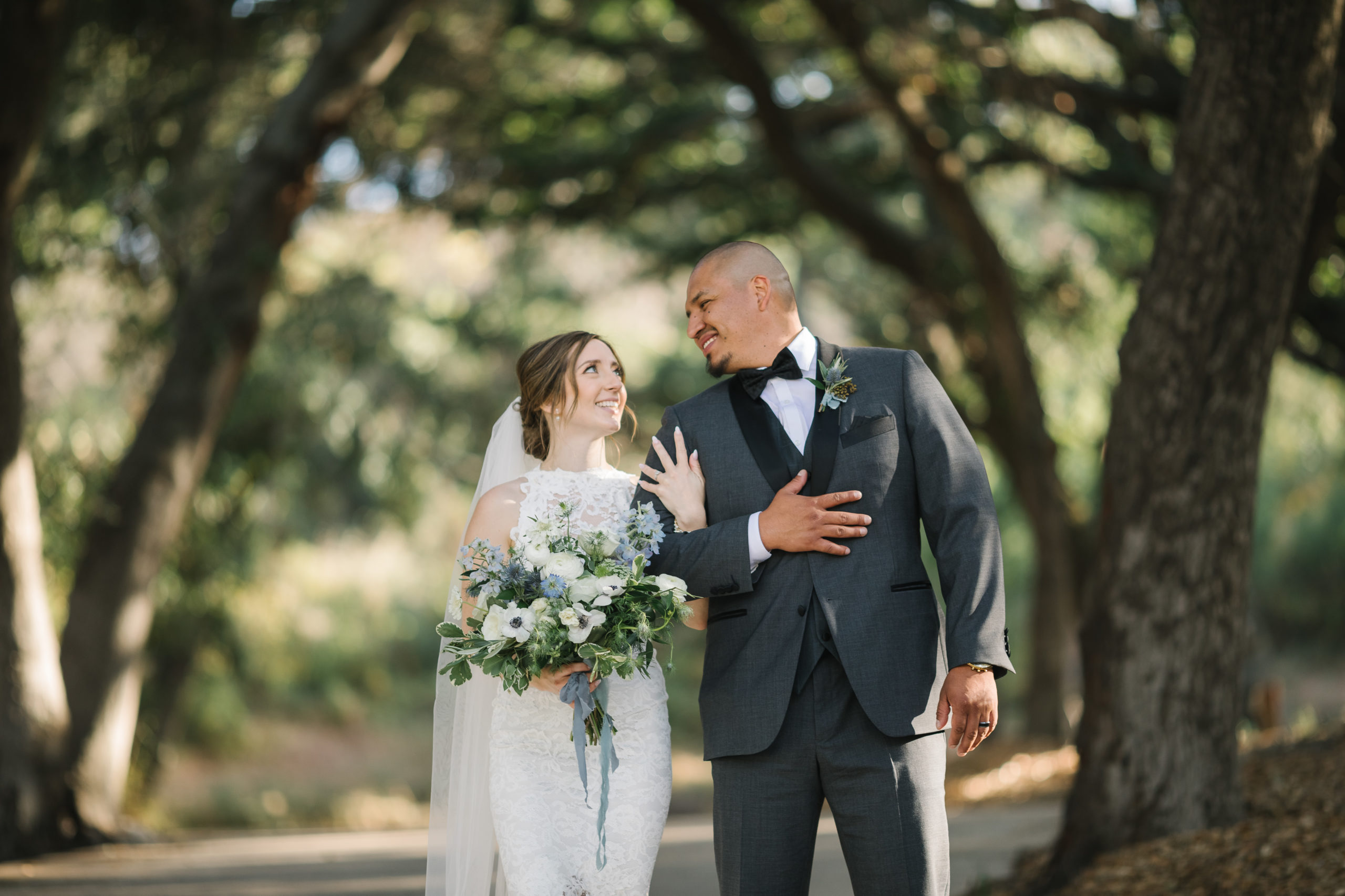 bride in high neckline lace wedding dress and white and blue bridal bouquet holds the arm of groom in dark grey suit and black bowtie
