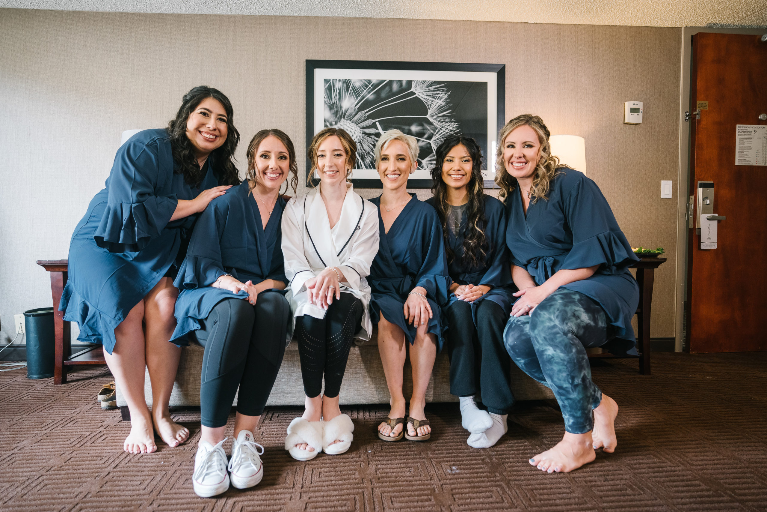 bride to be in white robe sits with bridesmaids in dark blue silk robes