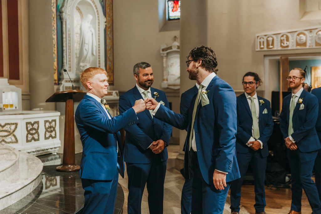 groom in navy blue suite and green bowtie fist bumps groomsmen in navy suite and green tie at altar