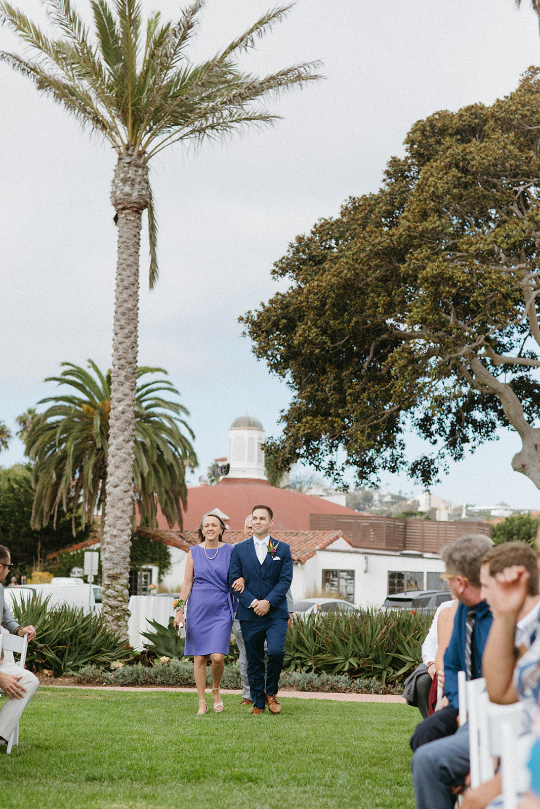 groom in navy blue suit and white tie walks with mom down the aisle
