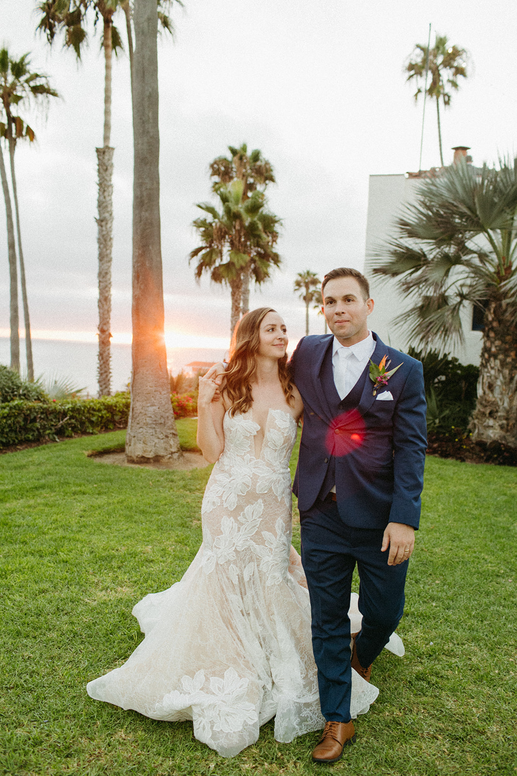 bride in strapless champagne with lace overlay mermaid wedding dress and tropical bouquet with groom in blue suit with white tie take sunset portrait shot overlooking the ocean