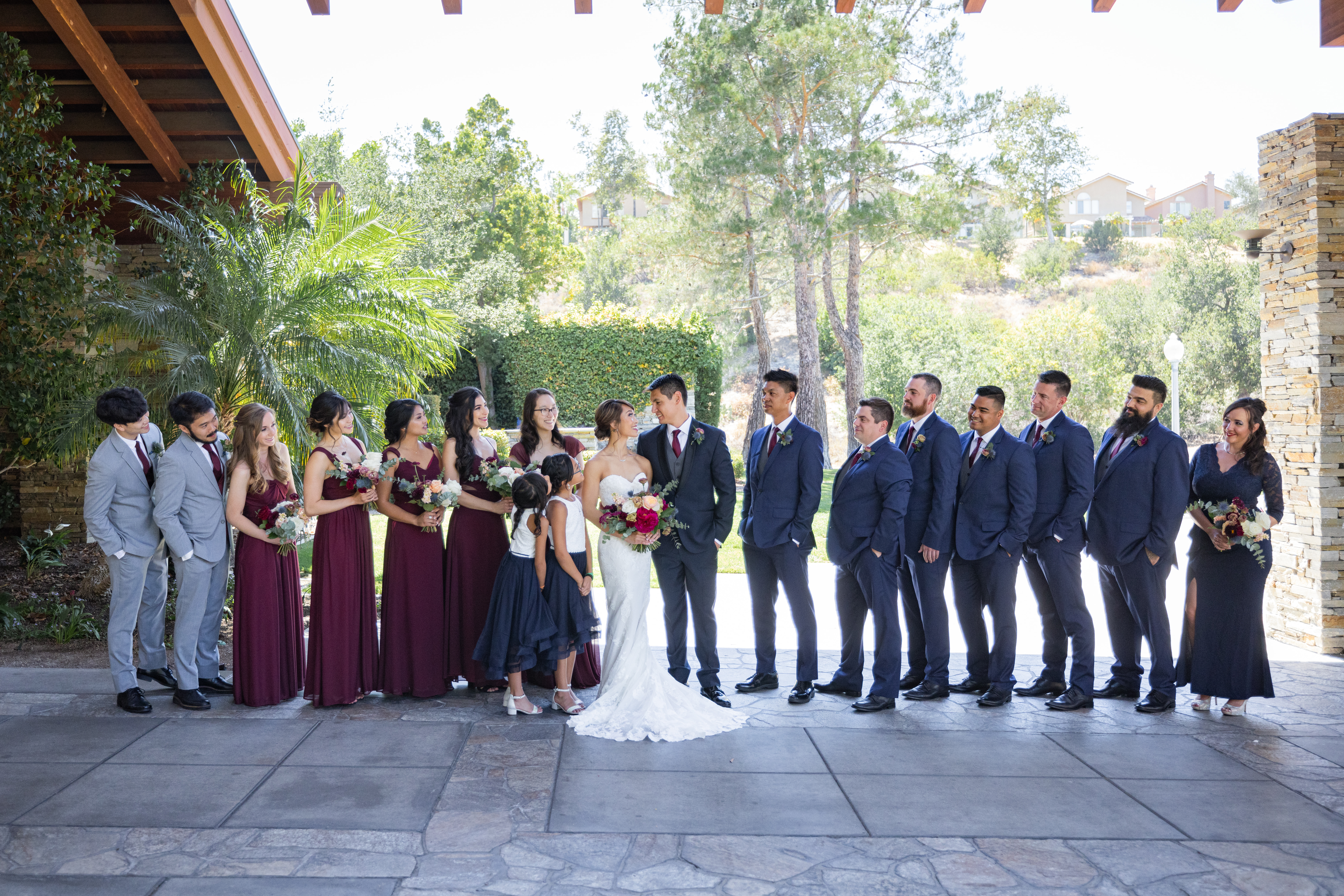 bride in strapless wedding dress and jewel toned bridal bouquet and groom in navy tuxedo suit with black lapels, grey vest and maroon tie stand with co-ed wedding party