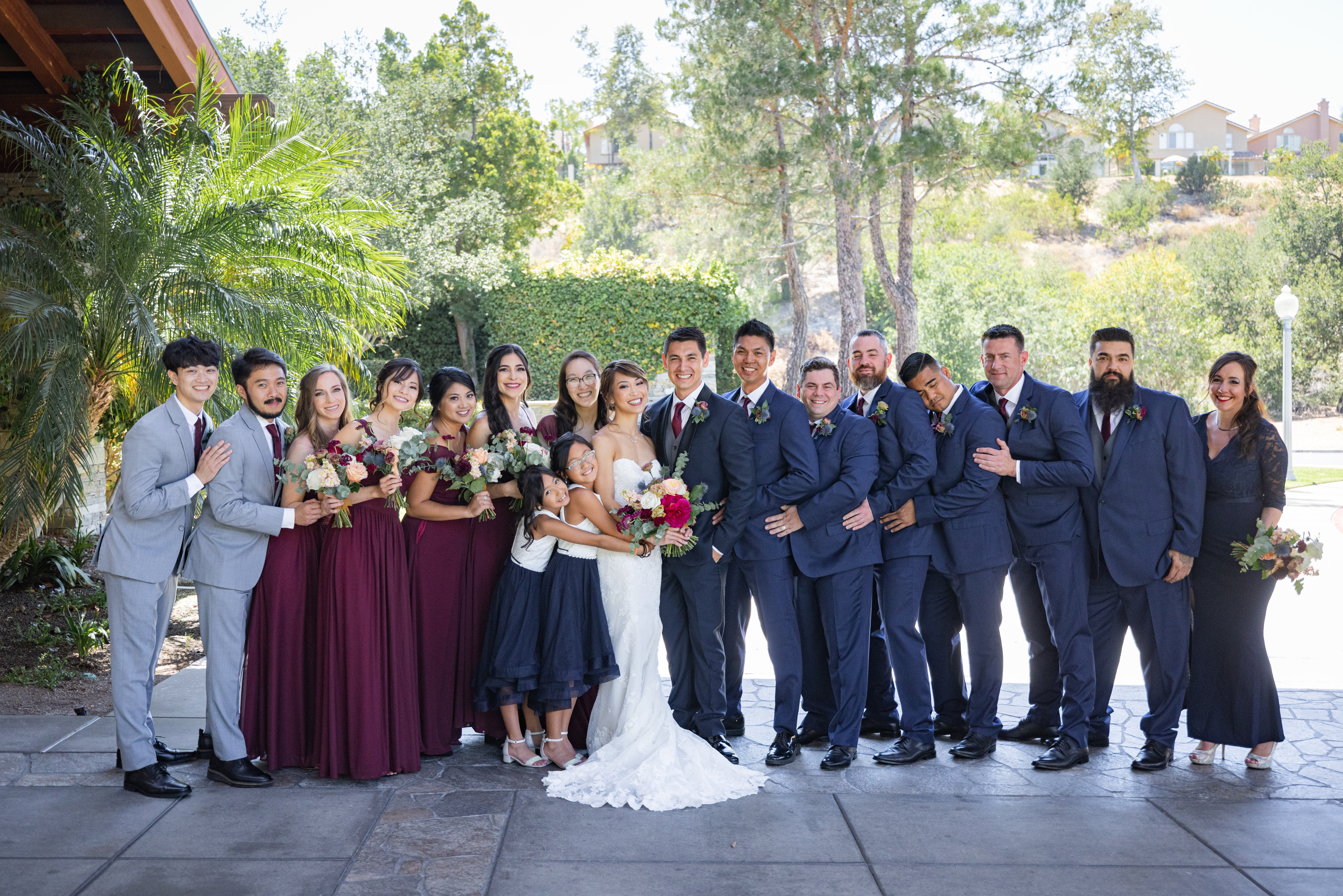 bride in strapless wedding dress and jewel toned bridal bouquet and groom in navy tuxedo suit with black lapels, grey vest and maroon tie stand with co-ed wedding party
