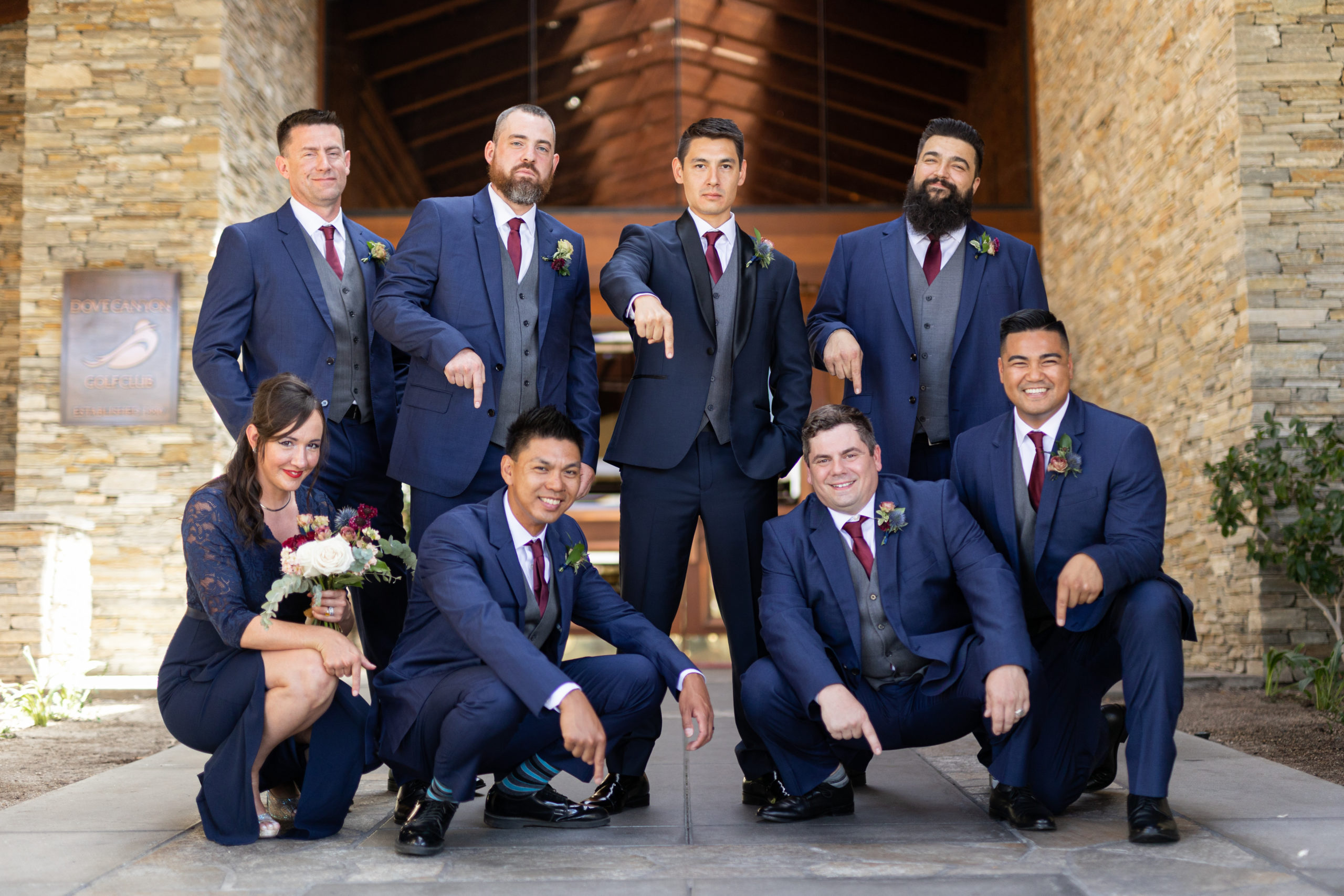 groom in navy tuxedo suit with black lapels, grey vest and maroon tie stand with co-ed wedding party in navy