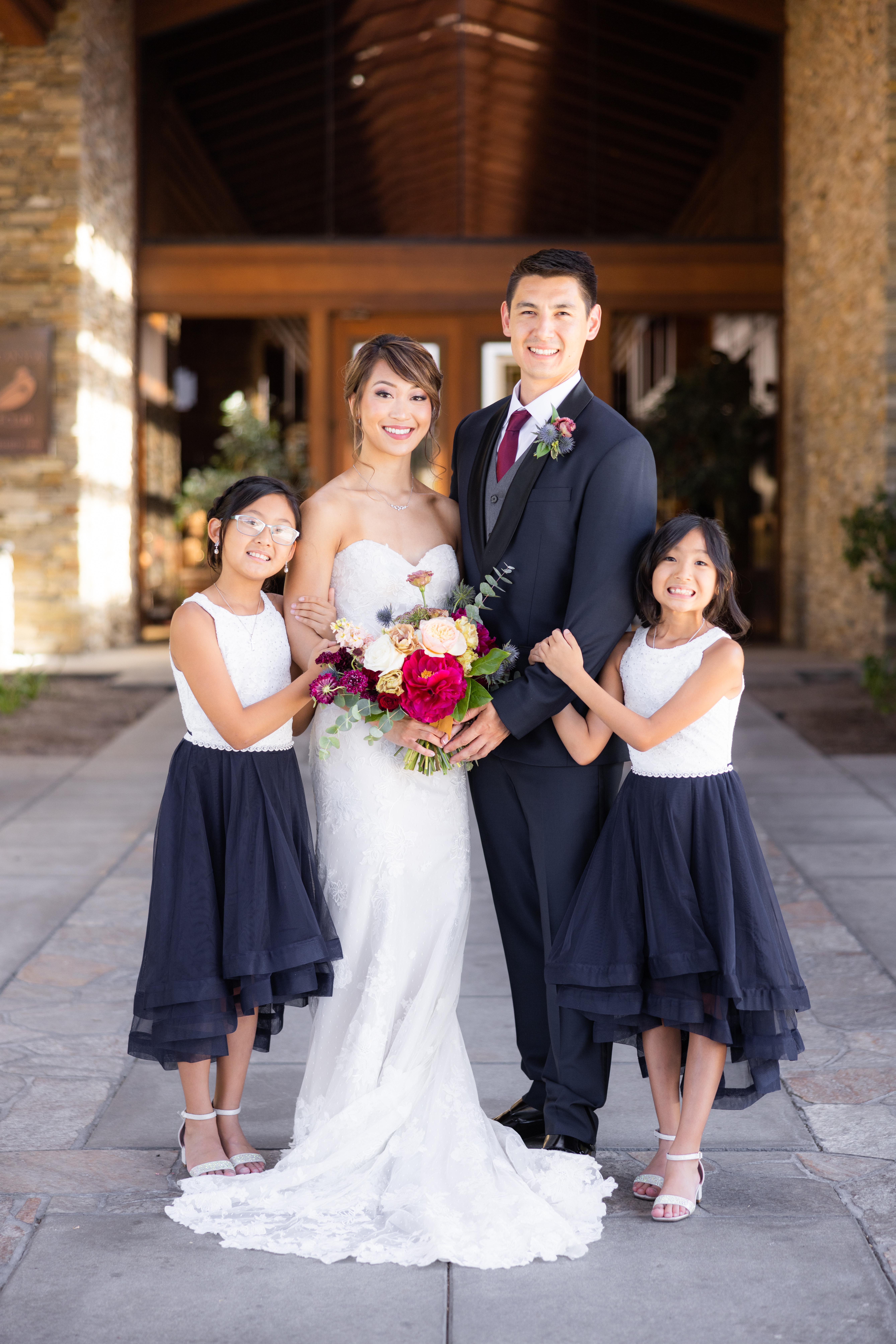 bride in strapless lace wedding dress and groom in navy tuxedo stand with flower girls in coordinating navy and white dresses