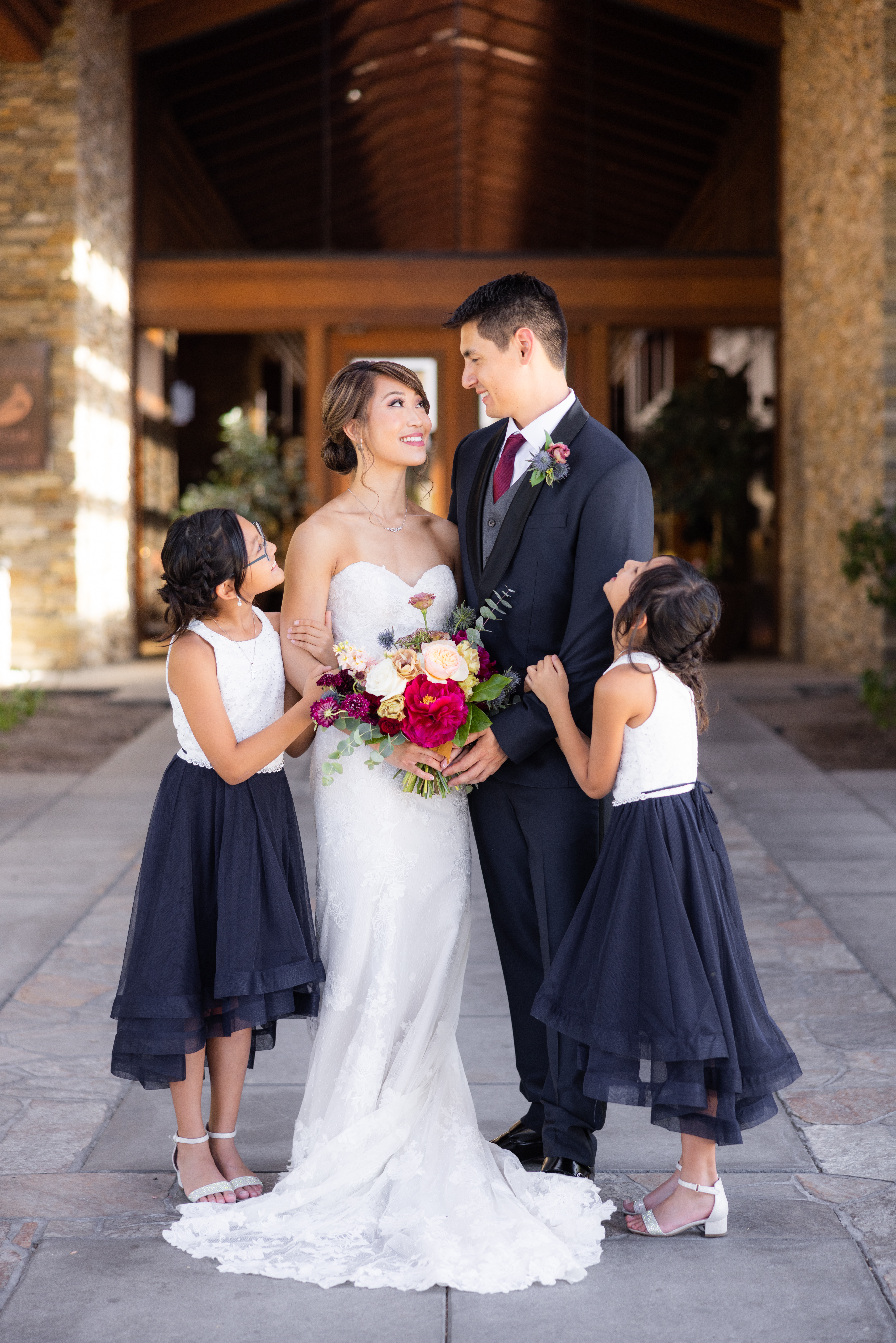 bride in strapless lace wedding dress and groom in navy tuxedo stand with flower girls in coordinating navy and white dresses