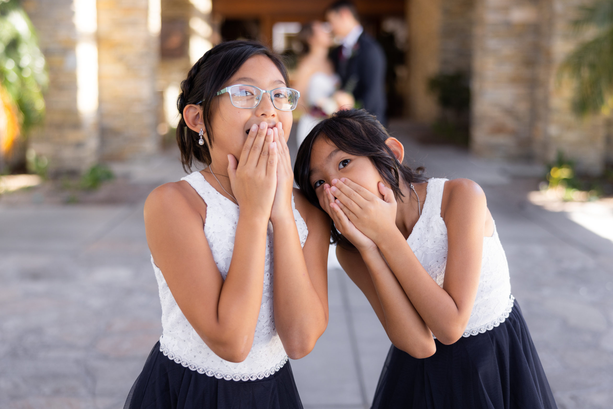 flower girls in white and navy dresses cover their mouths in surprise as bride and groom kiss in the background