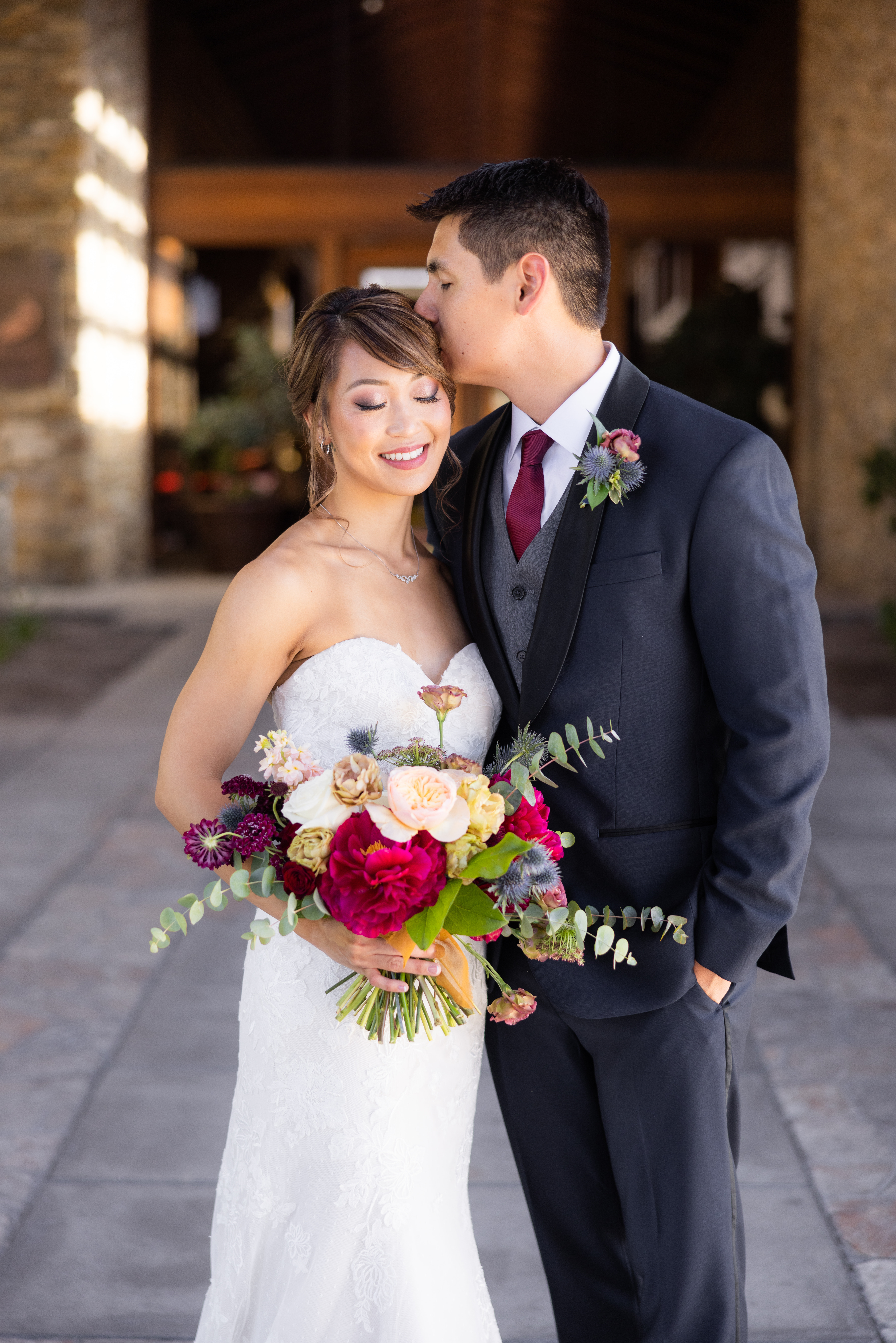 bride in strapless lace wedding dress with jewel toned bridal bouquet stands with groom in navy suit with black lapels, grey vest and maroon tie