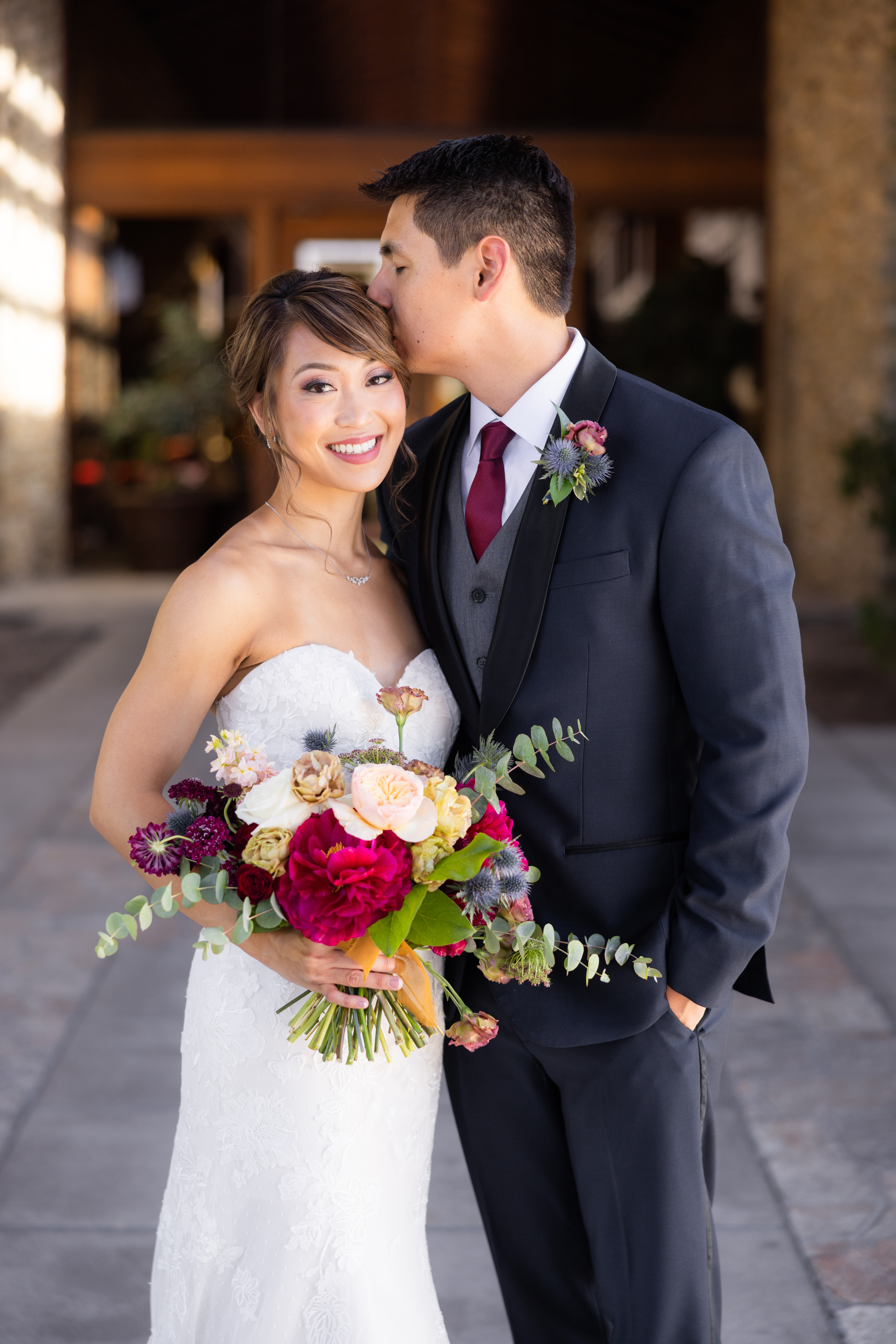 bride in strapless lace wedding dress with jewel toned bridal bouquet stands with groom in navy suit with black lapels, grey vest and maroon tie