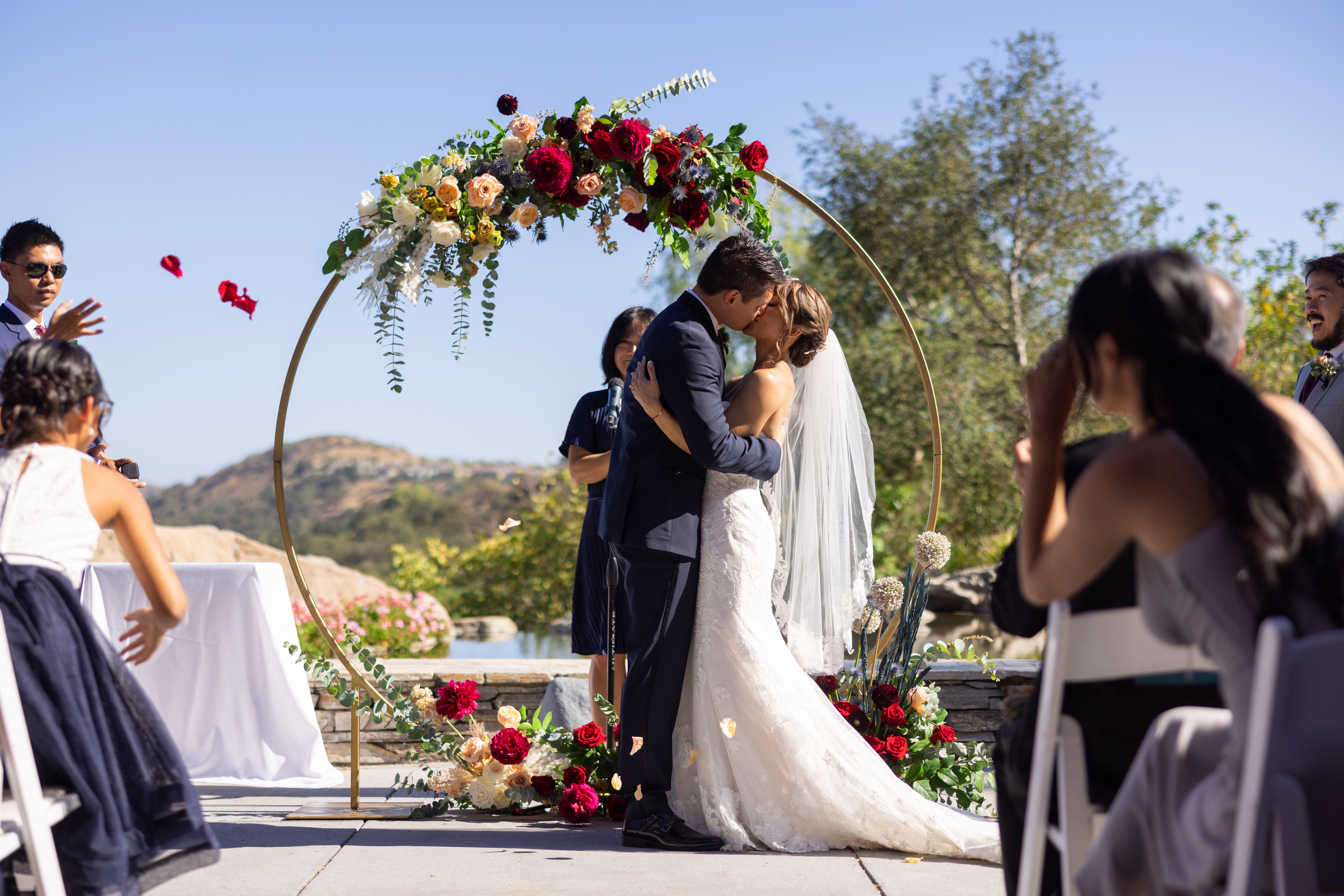bride and groom first kiss during outdoor wedding ceremony at Dove Canyon Gold Club with with gold circle ceremony arch with jewel toned florals