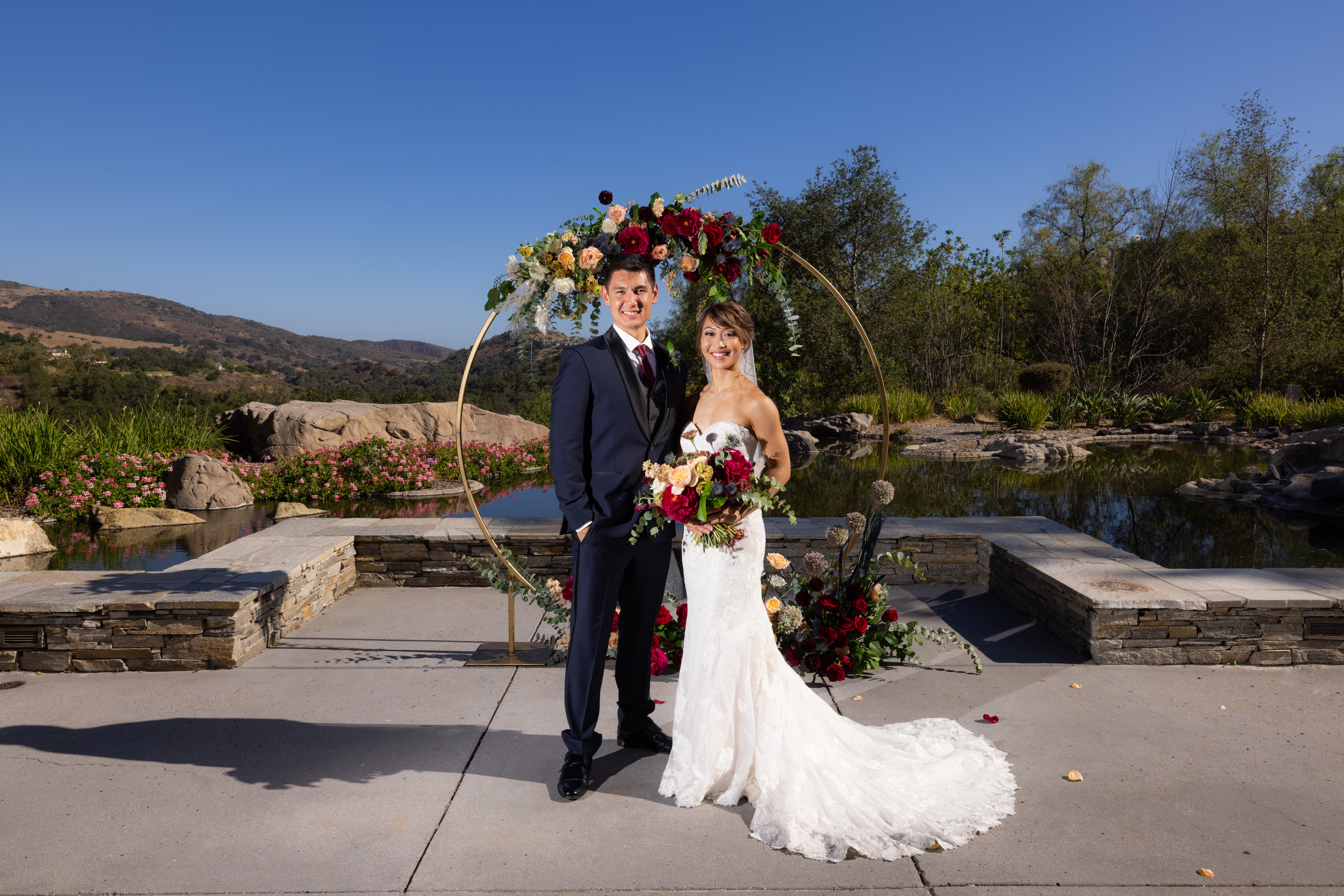 bride in strapless lace wedding dress and groom in navy tuxedo stand in front of gold circle ceremony arch with jewel toned florals