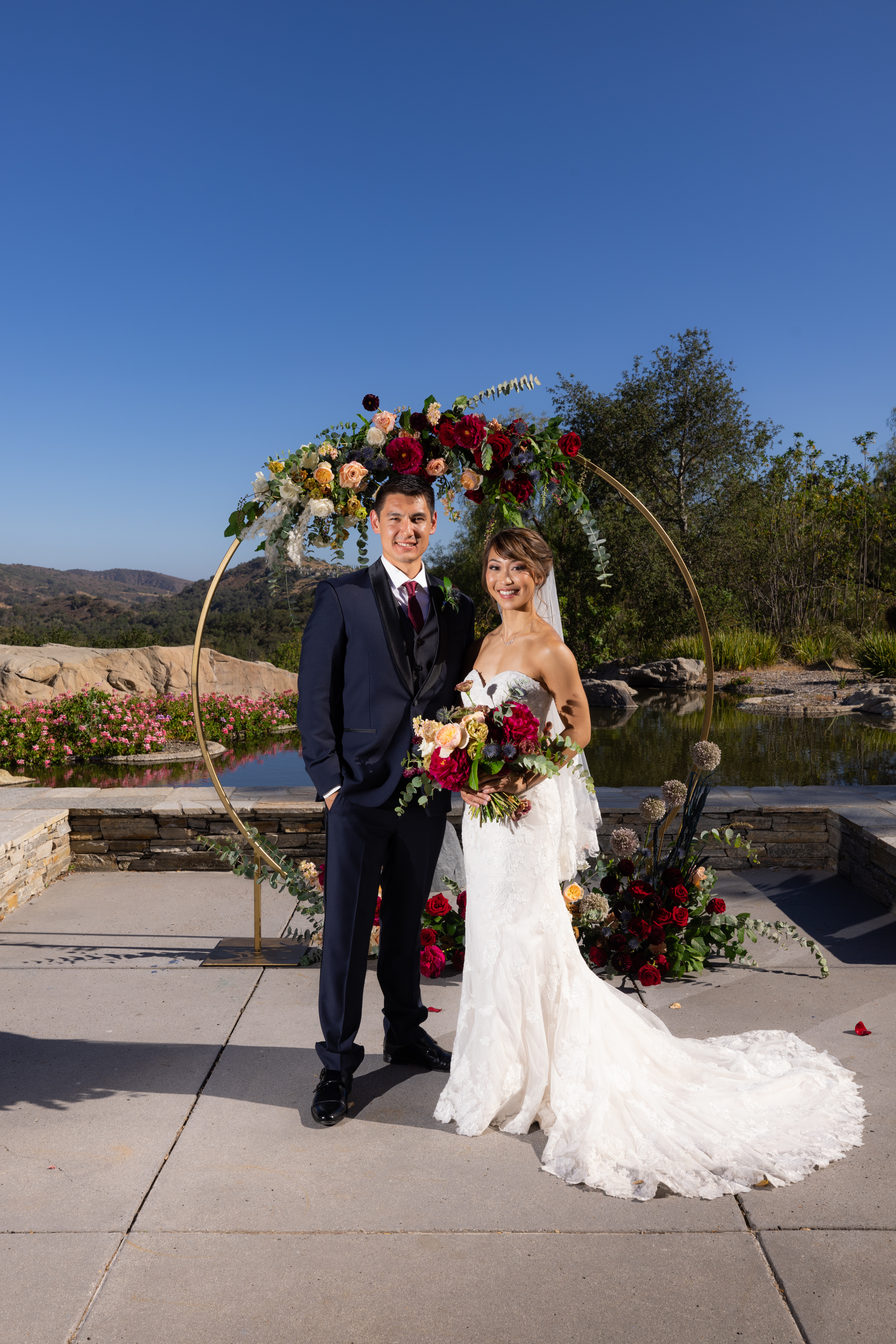 bride in strapless lace wedding dress and groom in navy tuxedo stand in front of gold circle ceremony arch with jewel toned florals
