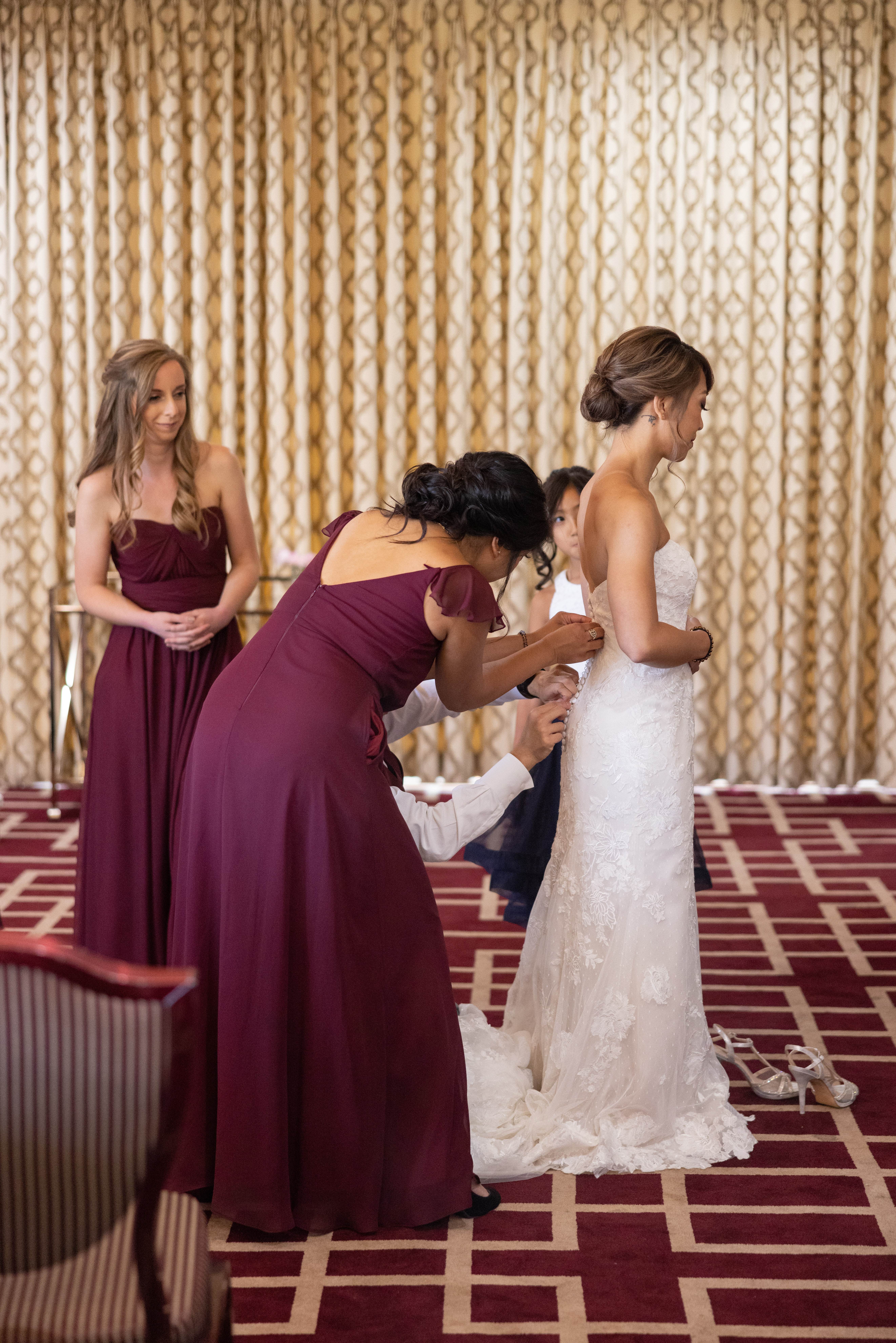 bride getting in strapless lace wedding dress with the help of her bridesmaids in jewel toned maroon dresses