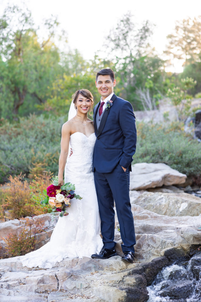 bride in strapless wedding dress with groom in dark blue suit and maroon tie stand by a waterfall during sunset portrait shots