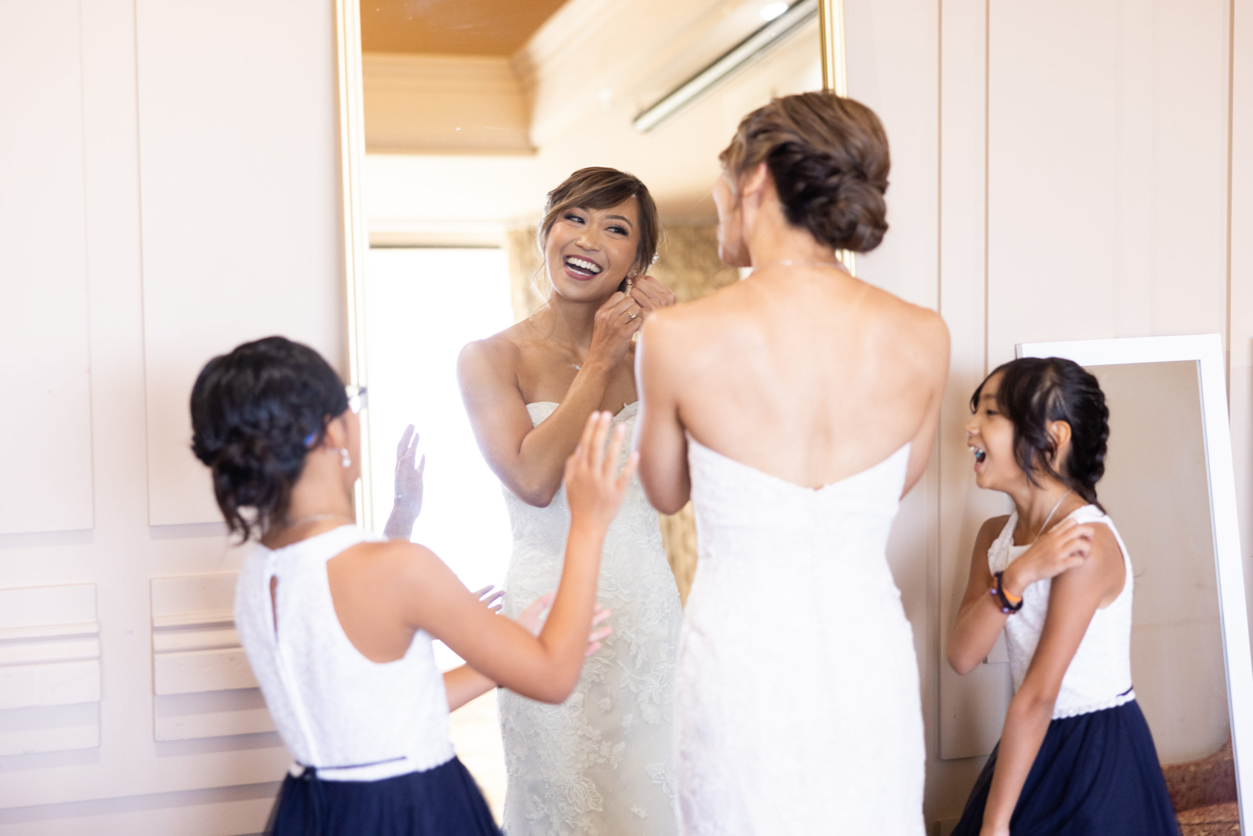 bride in strapless lace wedding dress putting in jewelry in mirror with help from flower girls in navy blue skirts
