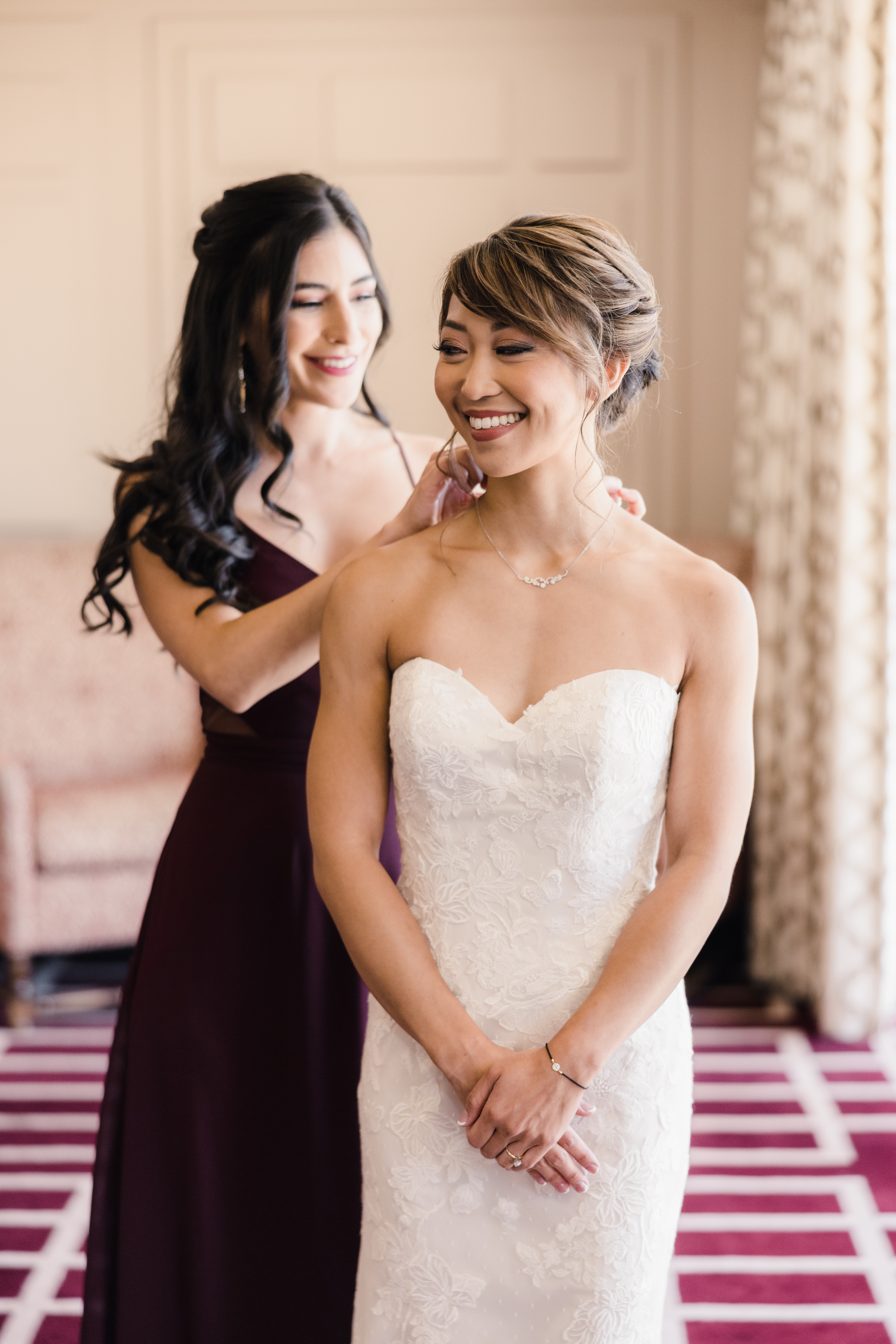 bride in strapless lace wedding dress and chignon hairstyle get help with jewelry from bridesmaid in jewel toned maroon dress