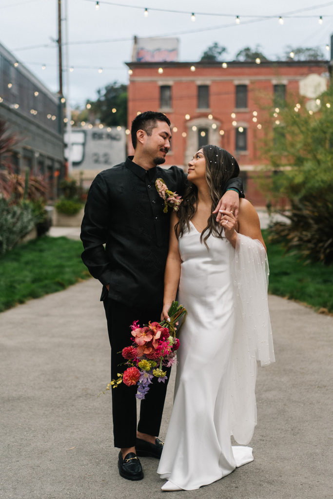 bride in modern silk white wedding dress with pearls in her hair holding bright bridal bouquet stands with groom in all black suit outside The Revery with market lights behind them