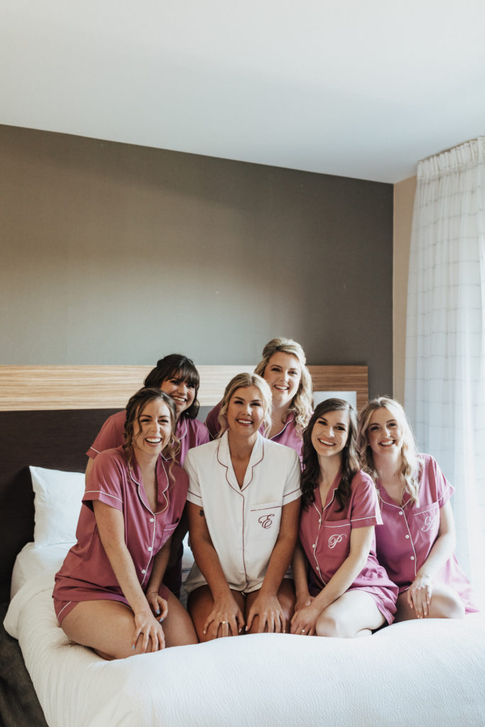 bride in satin pjs with pink trim sits on bed with bridesmaids in satin pink pjs with white trim
