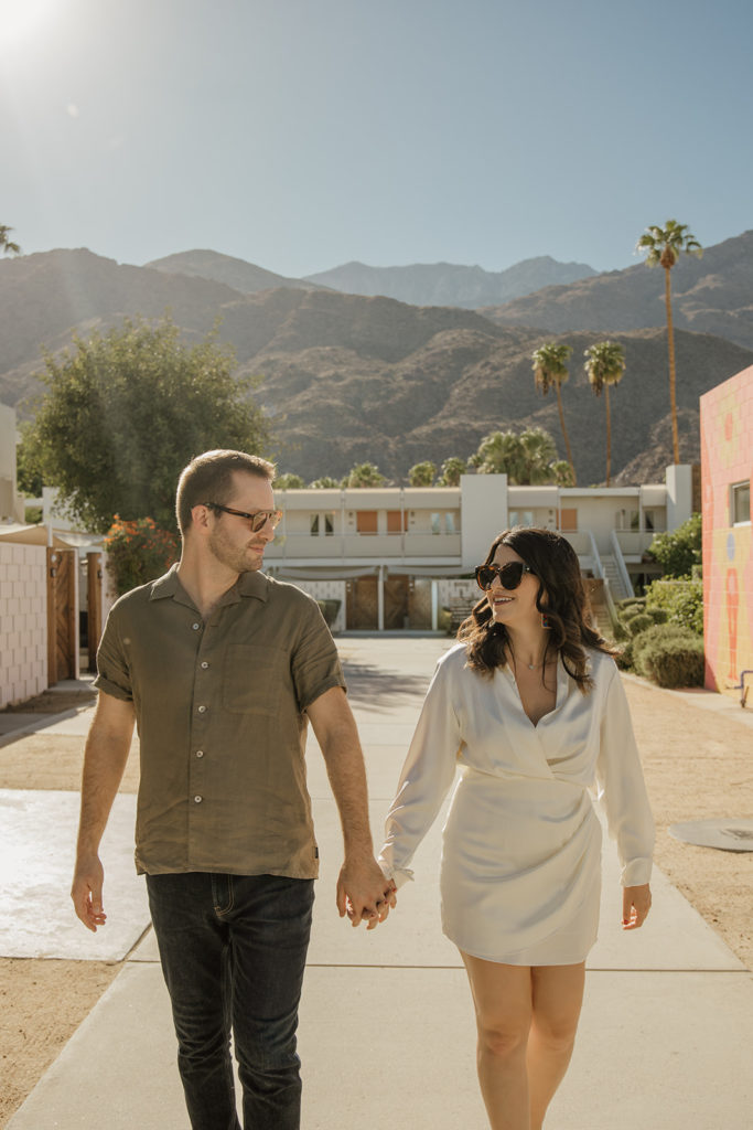 mid-century inspired desert engagement photo session with bride to be in silk mini dress and groom in brown button up walking with mountains in background