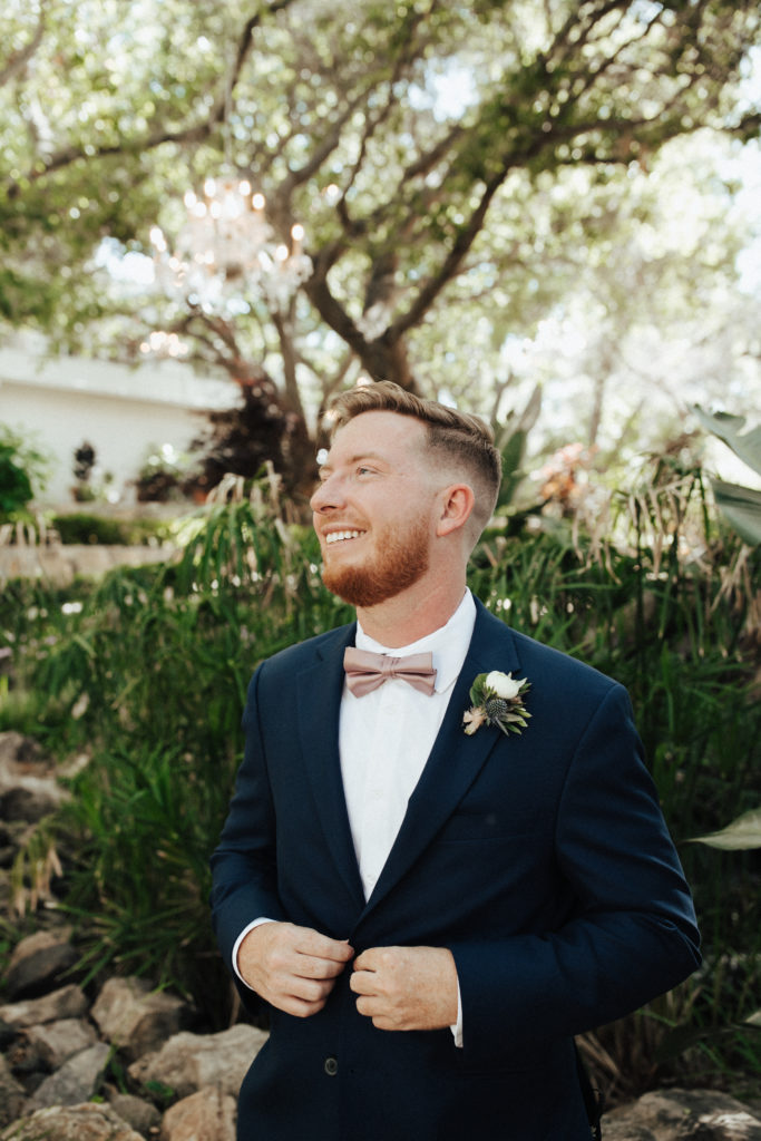 groom in black suit with soft pink bow tie stands in garden