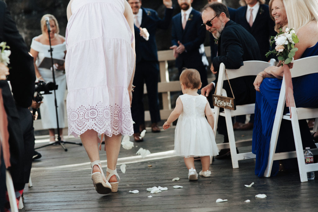 toddler flower girl walks down ceremony aisle in white dress