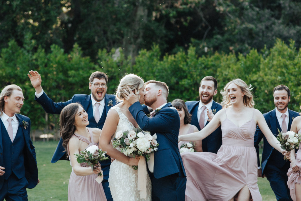 bride in deep vneck wedding dress with groom in black suit with pink tie stands with wedding party in pink bridesmaid dresses and black suits with pink ties during spring wedding at Calamigos Ranch