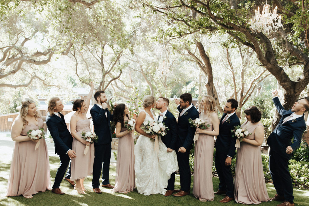 bride in deep vneck wedding dress with groom in black suit with pink tie stands with wedding party in pink bridesmaid dresses and black suits with pink ties during spring wedding at Calamigos Ranch