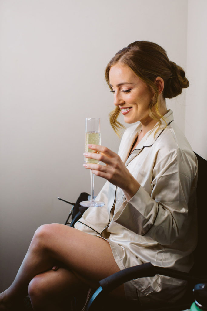 bride with glass of champagne in beige silk pajamas getting hair and makeup done 