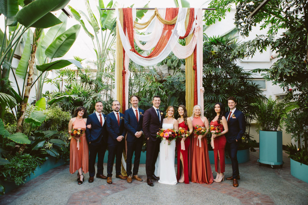 bride and groom stand with co-ed wedding party in front of earth toned ceremony drapery 