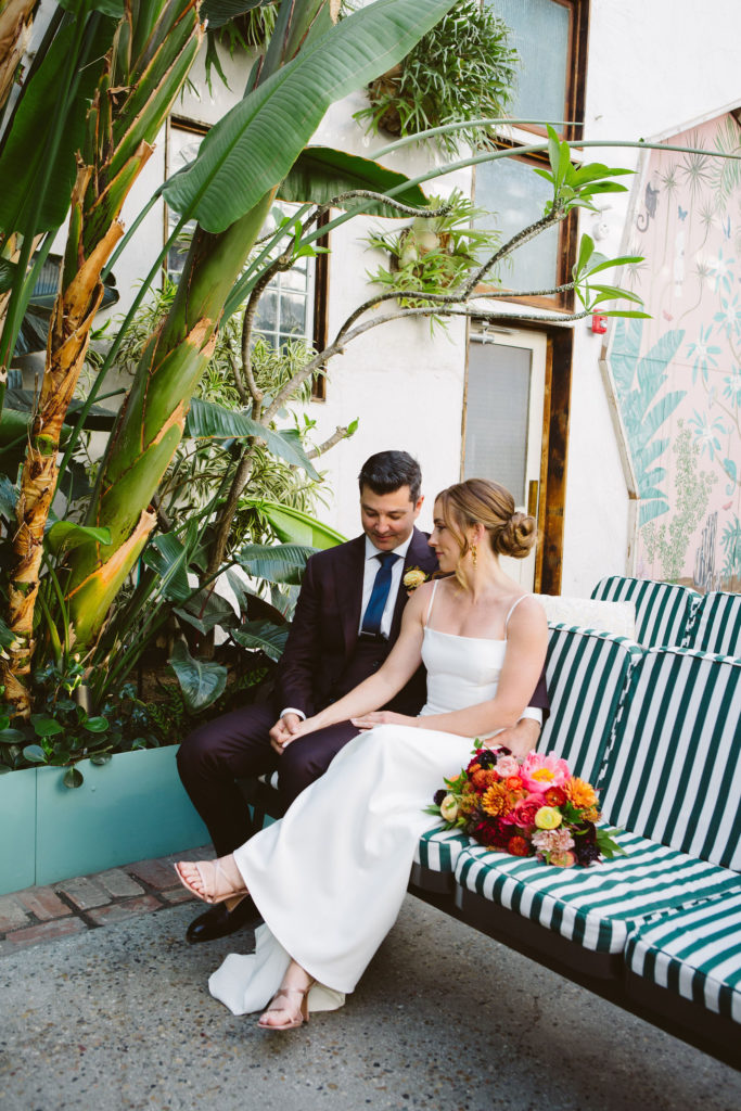 bride in modern square neck spaghetti strap wedding dress with groom in plum suit with blue tie sit in green striped chairs