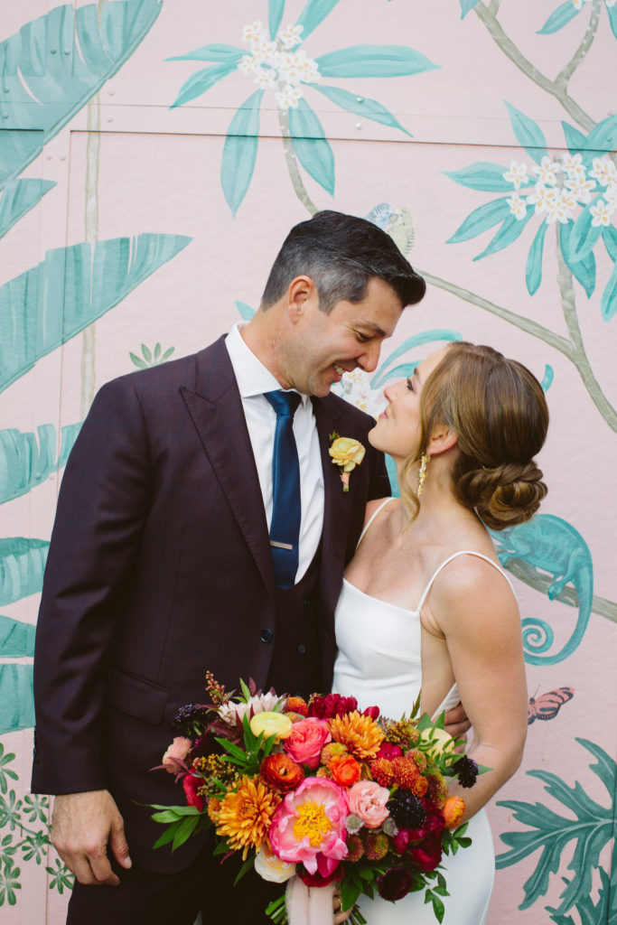 bride in modern square neck spaghetti strap wedding dress holding colorful bouquet with groom in deep plum suit and blue tie stand in front of pink keyhole wall at Valentine DTLA