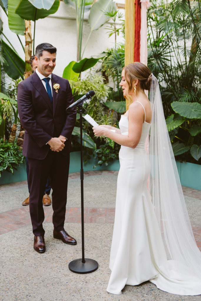 bride in modern square neck wedding dress with spaghetti straps and cathedral veil reads vows to groom in plus suit with blue tie during wedding ceremony at Valentine DTLA