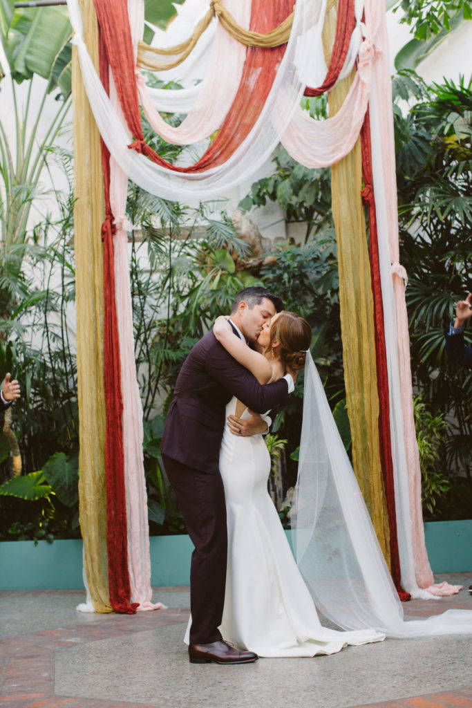 first kiss with bride in modern square neck wedding dress with spaghetti straps and cathedral veil and groom in plum suit in front of earth toned drapery for ceremony arch during wedding ceremony at Valentine DTLA
