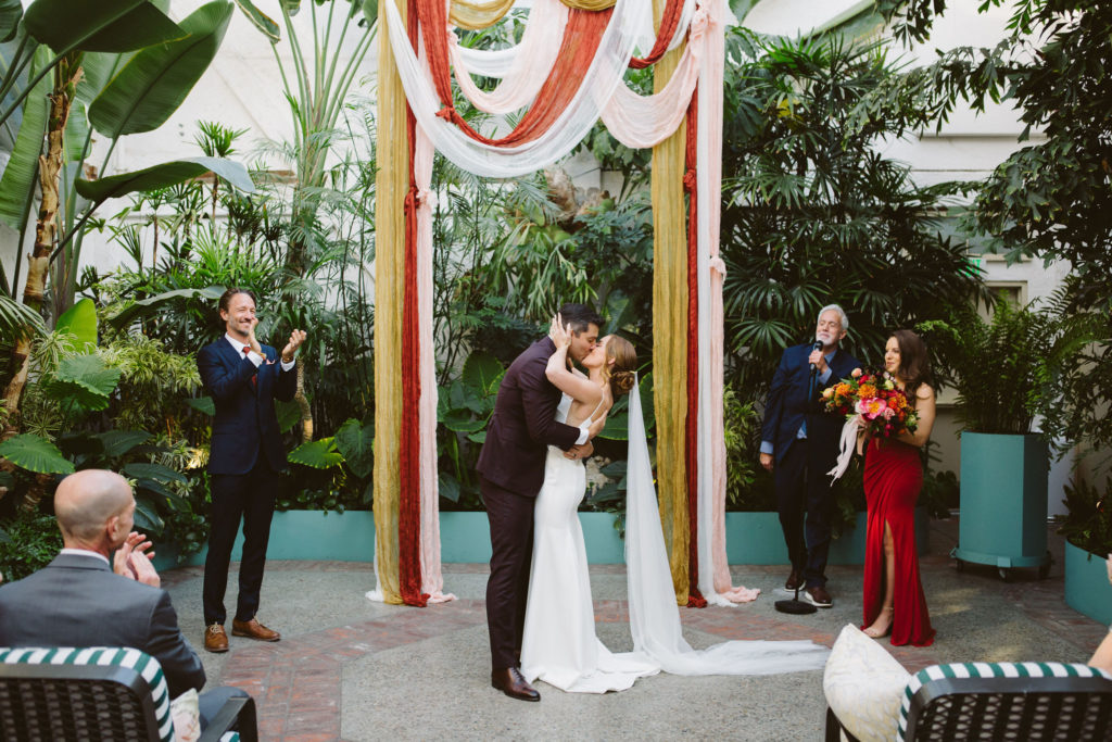 first kiss with bride in modern square neck wedding dress with spaghetti straps and cathedral veil and groom in plum suit in front of earth toned drapery for ceremony arch during wedding ceremony at Valentine DTLA