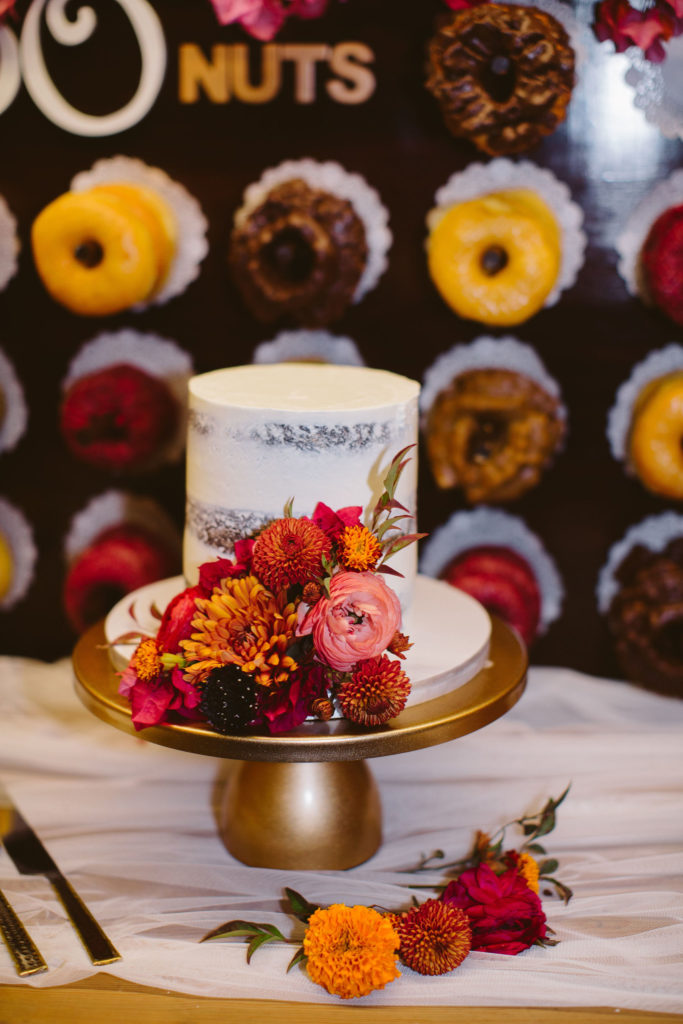 small single tier wedding cake with earth toned florals in front of a donut wall 
