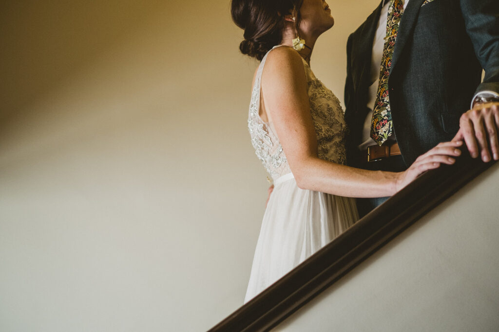 bride in embellished deep v neck wedding dress with a chiffon skirt and groom in in custom green suit take photos in stairwell of their hotel 