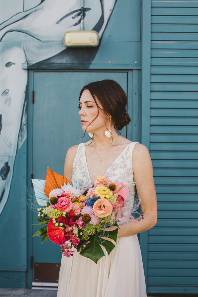 bride in embellished deep v neck wedding dress with a chiffon skirt holding rainbow colored wild jungle themed bouquet