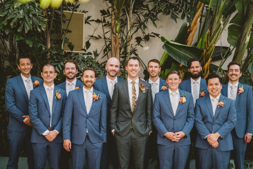 groom in custom suit and bright floral ties stands with groomsmen in blue suits and white ties 
